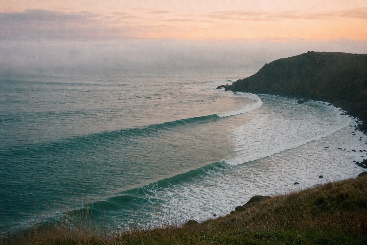 Empty lines of swell wrapping around Carrach Point at dawn, fog sitting on a peach horizon